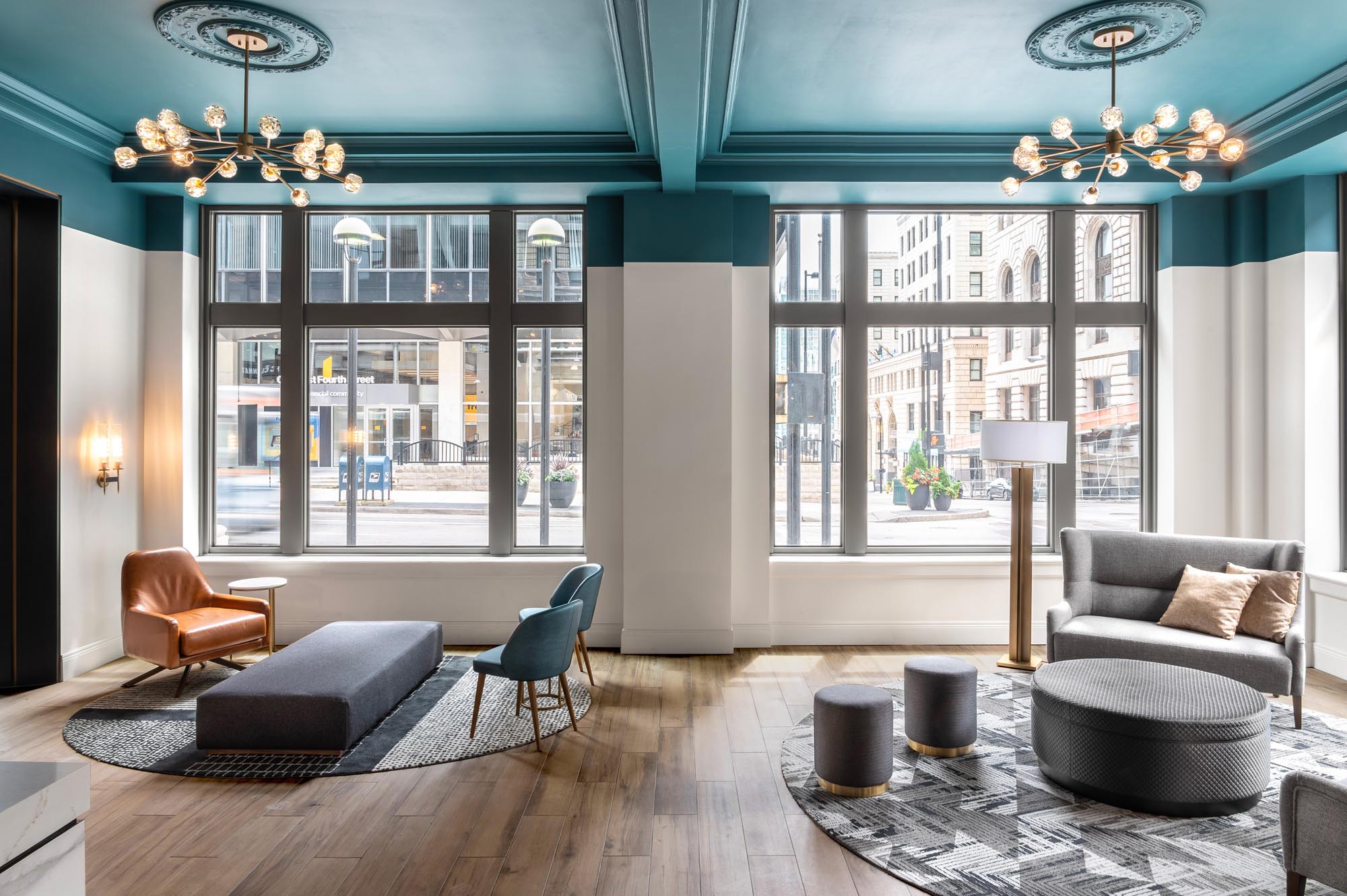 A lobby with large windows, stylish furniture, and city views. Light-colored walls contrast with teal accents. Seating includes a grey sofa, leather armchair, and teal chairs. Elegant lighting fixtures illuminate the space.